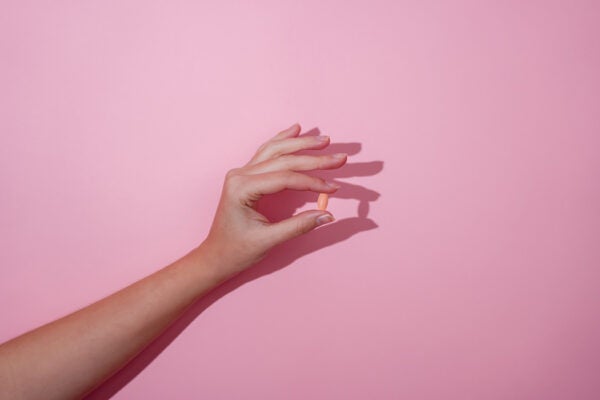 Hand holding a single pill between fingers on a pink background